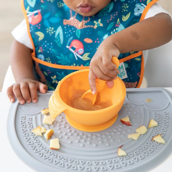 A child in a bib uses Bumkins Silicone First Feeding Set: Tangerine to self-feed with an orange spoon and bowl on the highchair tray.