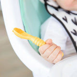 Close-up of a baby self-feeding with the Bumkins Silicone First Feeding Set in Tangerine, sitting in a high chair, wearing a bib.