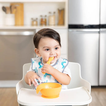 Toddler self-feeding with Bumkins Silicone First Feeding Set in Tangerine, wearing a bib, holding a bowl and spoon.