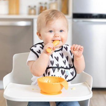 A baby in a high chair uses the Bumkins Silicone First Feeding Set: Tangerine, sporting a heart-patterned bib for self-feeding.