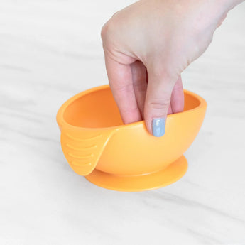 Hand lifts a Bumkins Silicone First Feeding Set: Tangerine bowl from a white marble surface.