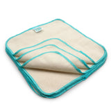 Cream-colored napkin from Bumkins, 100% cotton with teal edging, folded to show layers, includes a corner tag.