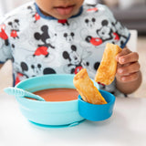 A child wearing a Mickey Mouse shirt enjoys soup and breadsticks from a Bumkins Silicone Grip Bowl: Blue at the white table.