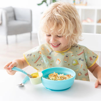 A smiling blonde child self-feeds from a Bumkins Silicone Grip Bowl: Blue, wearing a colorful bib and holding a spoon.