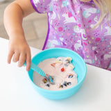 A child eats yogurt and fruit from a Bumkins Blue Silicone Grip Bowl, wearing a purple unicorn shirt, embracing toddler self-feeding.