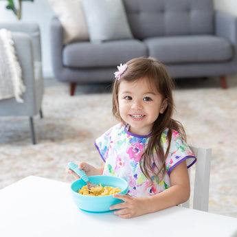 A cheerful child sits, mastering self-feeding with Bumkins Silicone Grip Bowl: Blue, spoon in hand, while the sofa is cozy in the background.