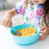 A toddler self-feeds mac and cheese from a Bumkins Silicone Grip Bowl: Blue, while wearing a vibrant bib.