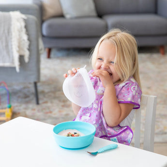 A child with long blonde hair in a purple shirt holds a Bumkins Silicone Grip Bowls lid near a Blue Suction Bowl at the table.