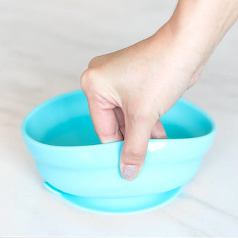 Hand holding a Bumkins Silicone Grip Bowl: Blue on marble, ideal for toddlers self-feeding with textured handle.