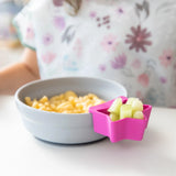 A Silicone Grip Bowl: Gray by Bumkins with macaroni and a pink dish of cucumbers sits on the table beside a floral shirt.