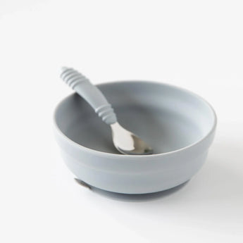 A Bumkins Silicone Grip Bowl in light gray and a matching spoon, both food-safe, rest on a white surface.