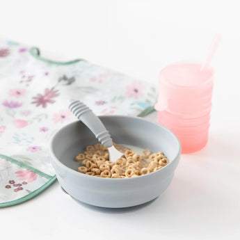 A Bumkins gray Silicone Grip Bowl, spoon, pink sippy cup, and floral bib sit on a white surface.