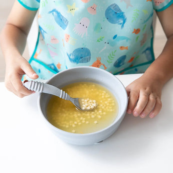 A child in an ocean bib holds a spoon over a Bumkins Silicone Grip Bowl: Gray, filled with soup and pasta, ready for their tasty adventure.