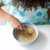 A child enjoys applesauce from a Bumkins Silicone Grip Bowl: Gray, spoon in hand, wearing a colorful animal-patterned shirt.