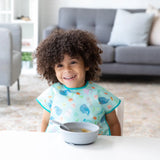 A curly-haired child in a colorful bib happily sits with a Bumkins gray Silicone Grip Bowl in the bright living room.