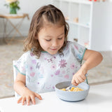 A young girl uses a spoon to eat cereal from the Bumkins Silicone Grip Bowl in gray, wearing a floral bib at a white table.