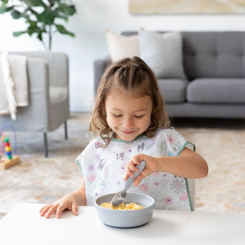 A young girl enjoys cereal in a Bumkins Silicone Grip Bowl: Gray, at the table with a cozy living room in the background.