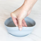 A hand gently grips a Bumkins Silicone Grip Bowl: Gray by its edge, fingers resting inside it against a light background.