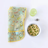 A vibrant bib, Bumkins Marble Silicone Grip Bowl of mac & peas, green cup, and toddler fork on a white surface.