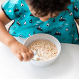 A child self-feeds cereal from a Bumkins Silicone Grip Bowl: Marble, wearing a blue bib with mountain and arrow patterns, holding a spoon.