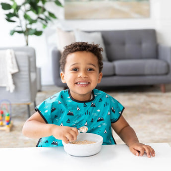 A child with a wide smile enjoys cereal from a Bumkins Silicone Grip Bowl: Marble, in a cozy living room with a sofa & plants nearby.