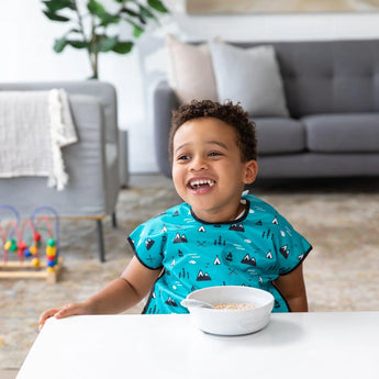 A child in a blue bib sits at the table mastering self-feeding with a Bumkins Silicone Grip Bowl: Marble, in a playful living room.