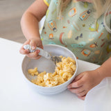 A child enjoys mac and cheese with a fork, wearing a colorful bib from Bumkins, using the Silicone Grip Bowl: Marble on the white table.