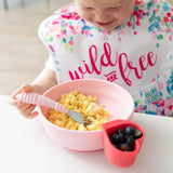 A child eats scrambled eggs from a pink Bumkins Silicone Grip Bowl with suction, using a pink fork. Berries await nearby.