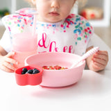 A toddler enjoys breakfast with Bumkins pink Silicone Grip Bowl, featuring suction base, plus a matching cup and utensil set.