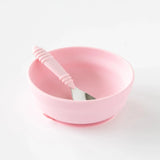 A Bumkins Silicone Grip Bowl in pink, featuring a spoon and interior ridge, sits elegantly on a white surface.