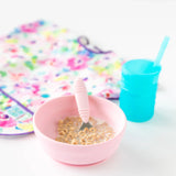 A Bumkins Silicone Grip Bowl: Pink with cereal and milk, beside a blue cup with a straw and a colorful bib.