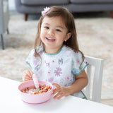 A smiling toddler with brown hair enjoys cereal from a Bumkins Silicone Grip Bowl in pink, wearing a floral bib and using a spoon.