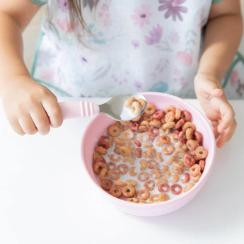 A child wearing a floral shirt enjoys colorful cereal and milk from Bumkins pink Silicone Grip Bowl, using a spoon.