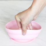 A hand squeezes a flexible Bumkins Silicone Grip Bowl: Pink with an interior ridge on a white surface.