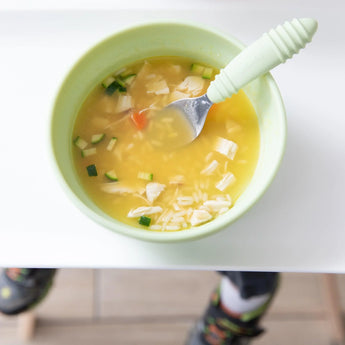 A Bumkins Silicone Grip Bowl: Sage with chicken, rice, and veggies sits on the table alongside a green-handled spoon; a figure lingers nearby.