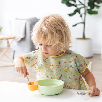 A young child with blonde hair uses a fork to eat from a Bumkins Silicone Grip Bowl in Sage, wearing a patterned bib in a bright room.