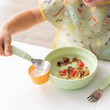 A child in a patterned bib scoops yogurt from a yellow cup onto a green plate with snacks, using Bumkins Sage Silicone Grip Bowl.