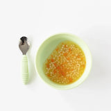 A Bumkins Silicone Grip Bowl in Sage, filled with soup and pasta, sits beside a green-handled spoon on a white background.