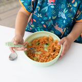 A child in a colorful shirt holds a fork over pasta at a white table with Bumkins Silicone Grip Bowl: Sage and spoon ready for use.