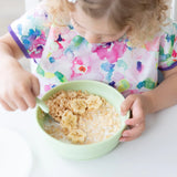 A child in a colorful floral shirt enjoys cereal with banana slices from a Bumkins Silicone Grip Bowl in Sage.