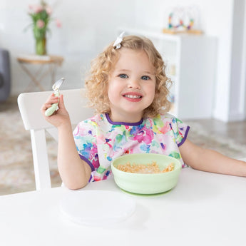 A curly-haired child beams enjoying cereal from a Bumkins Silicone Grip Bowl: Sage, adding charm with their bright top and bow.