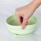 A hand lifts the Bumkins Silicone Grip Bowl in Sage with a strong suction base from a marble surface.