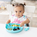 A toddler with pigtails enjoys a meal from a Bumkins Silicone Grip Dish: Blue, wearing a bib, with sections of fruits and veggies.