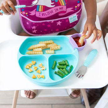 A child in a high chair enjoys crinkle-cut fries with ketchup on a blue Bumkins Silicone Grip Dish, plus green beans & cheese shapes.