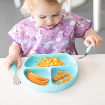 A toddler in a unicorn bib enjoys mac and cheese, peppers, and cubed food from a blue plate with Bumkins Spoon + Fork: Blue.