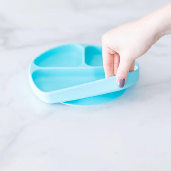 A hand lifts a Silicone Grip Dish: Blue by Bumkins from a marble surface, highlighting its firm suction base.