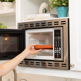 A hand places a Bumkins Silicone Grip Dish: Clay inside a stainless steel microwave, with white shelves and books in the background.