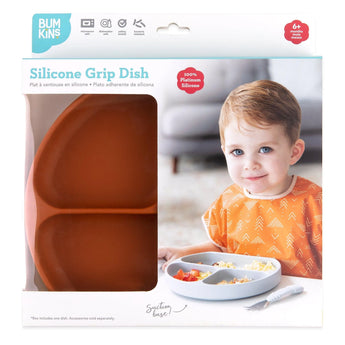 A child in a Bumkins Silicone Grip Dish: Clay, featuring a suction base and divided sections, wears an orange bib. Packaging is visible.