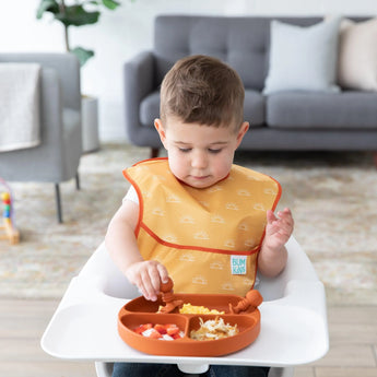 A toddler enjoys a meal from a Bumkins Silicone Grip Dish in Clay on the high chair, with a cozy gray sofa in the background.