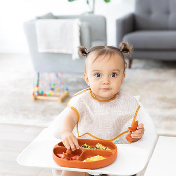 A toddler with pigtails enjoys a meal from a Bumkins Silicone Grip Dish: Clay, wearing a patterned bib in a cozy living room.